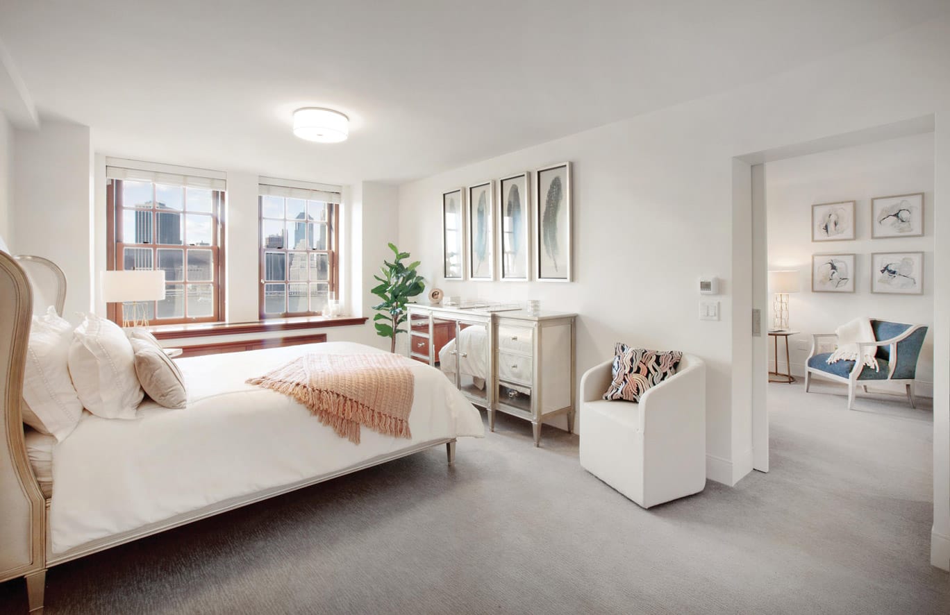 Brooklyn Heights Independent & Assisted Living Photo Gallery - Bedroom layout showcasing a neatly made bed, dresser, seating area, and a view of New York through mahogany framed windows with a view into the living area