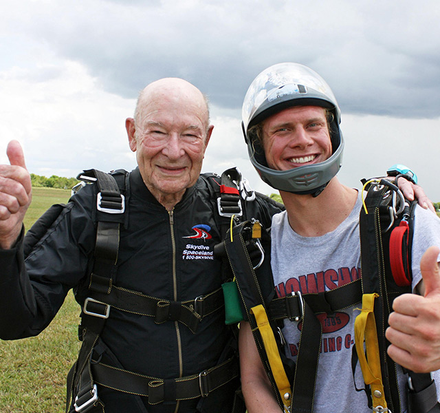 elderly gentleman and his son standing in a field wearing skydiving gear