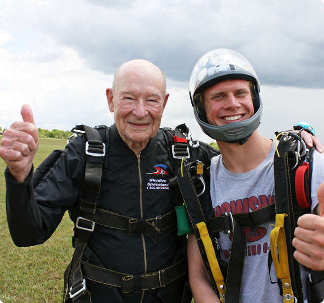 elderly gentleman and his son standing in a field wearing skydiving gear