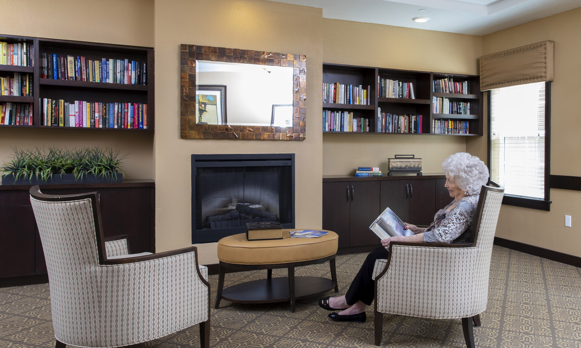 Lady reading a book in an armchair, facing a fireplace in in-laid bookshelfs