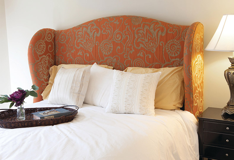 A bed with cream coloured duvet and a rust coloured headboard, night stand with antique lamp
