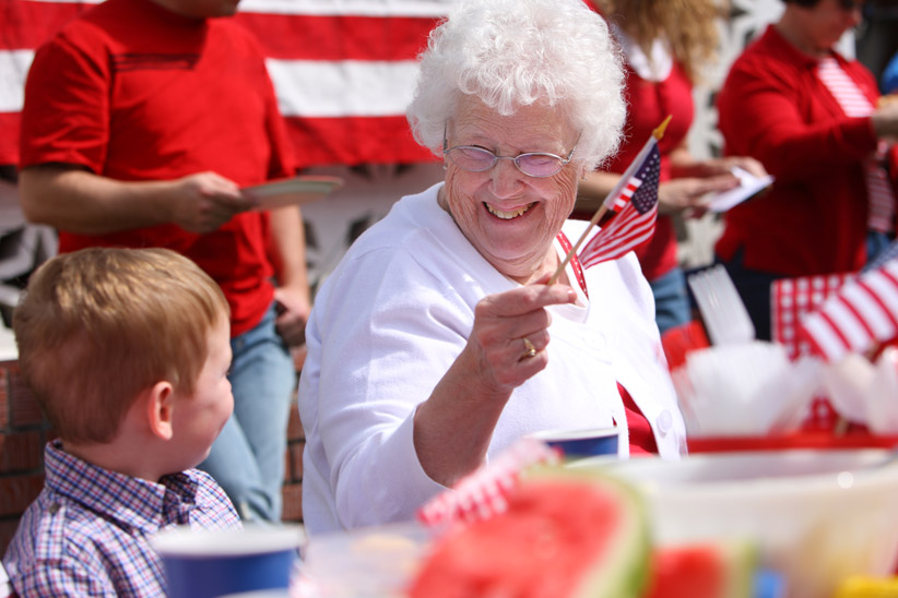 An elderly woman is holding a small American flag while gazing at her grandson and laughing