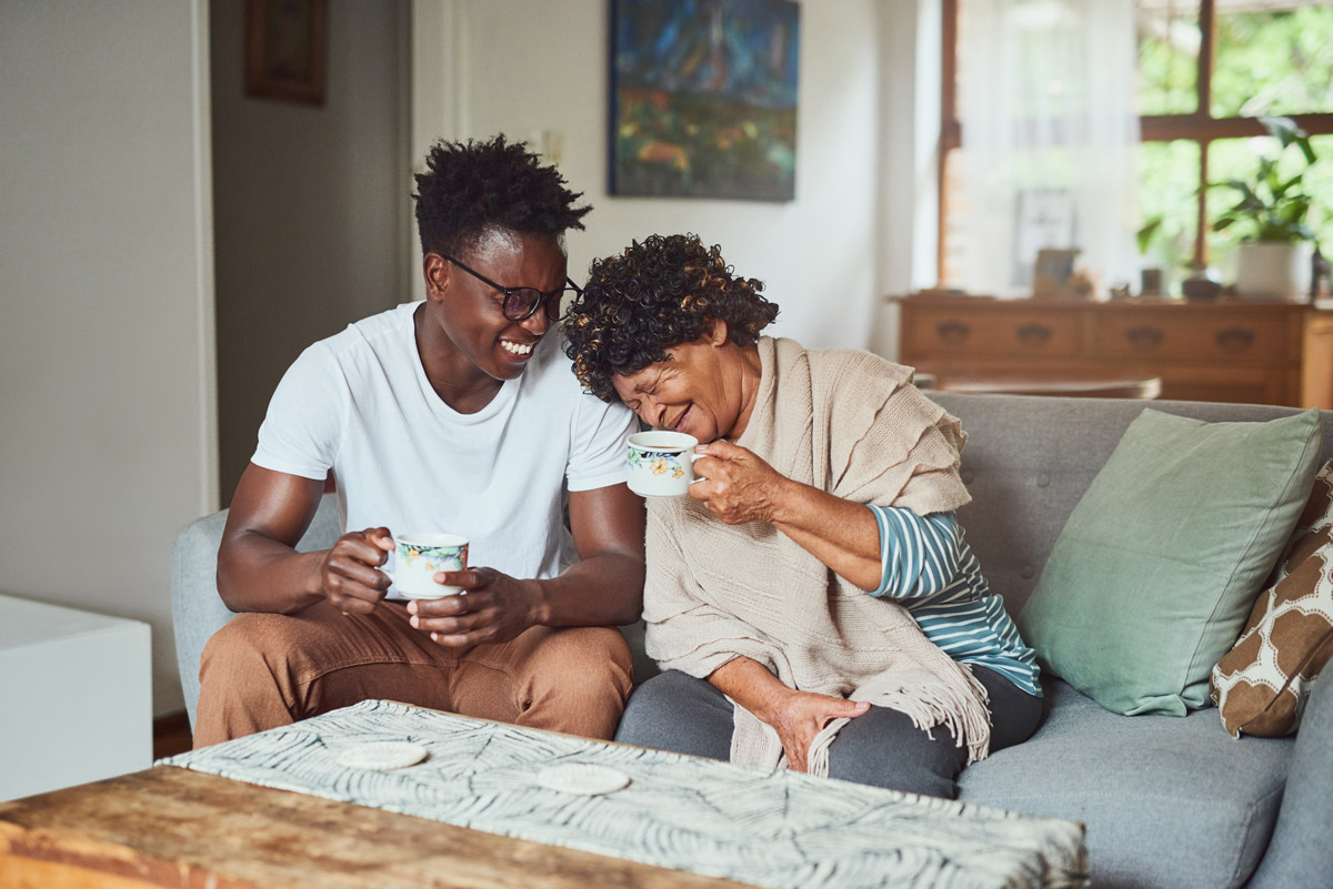 A young man and his elderly mother are sitting on a couch while holding their tea cups and laughing to one another