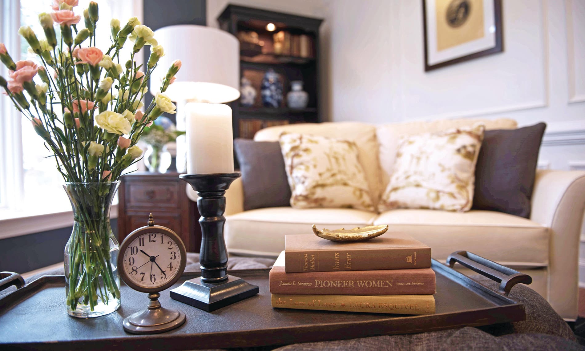 A vase featuring a colourful floral arrangements sits next to an analog clock, candle and several books with a white love seat and side table in the background. 