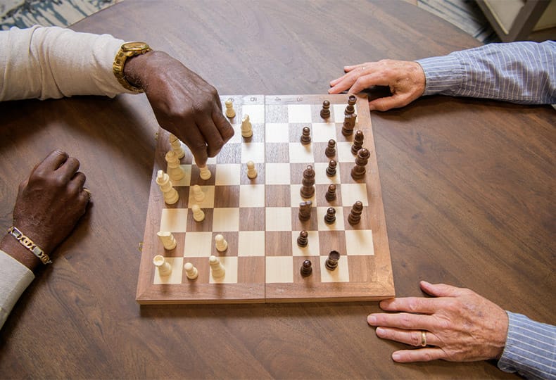 A pair of elderly gentleman's hands are visible on either side of a wooden chess board