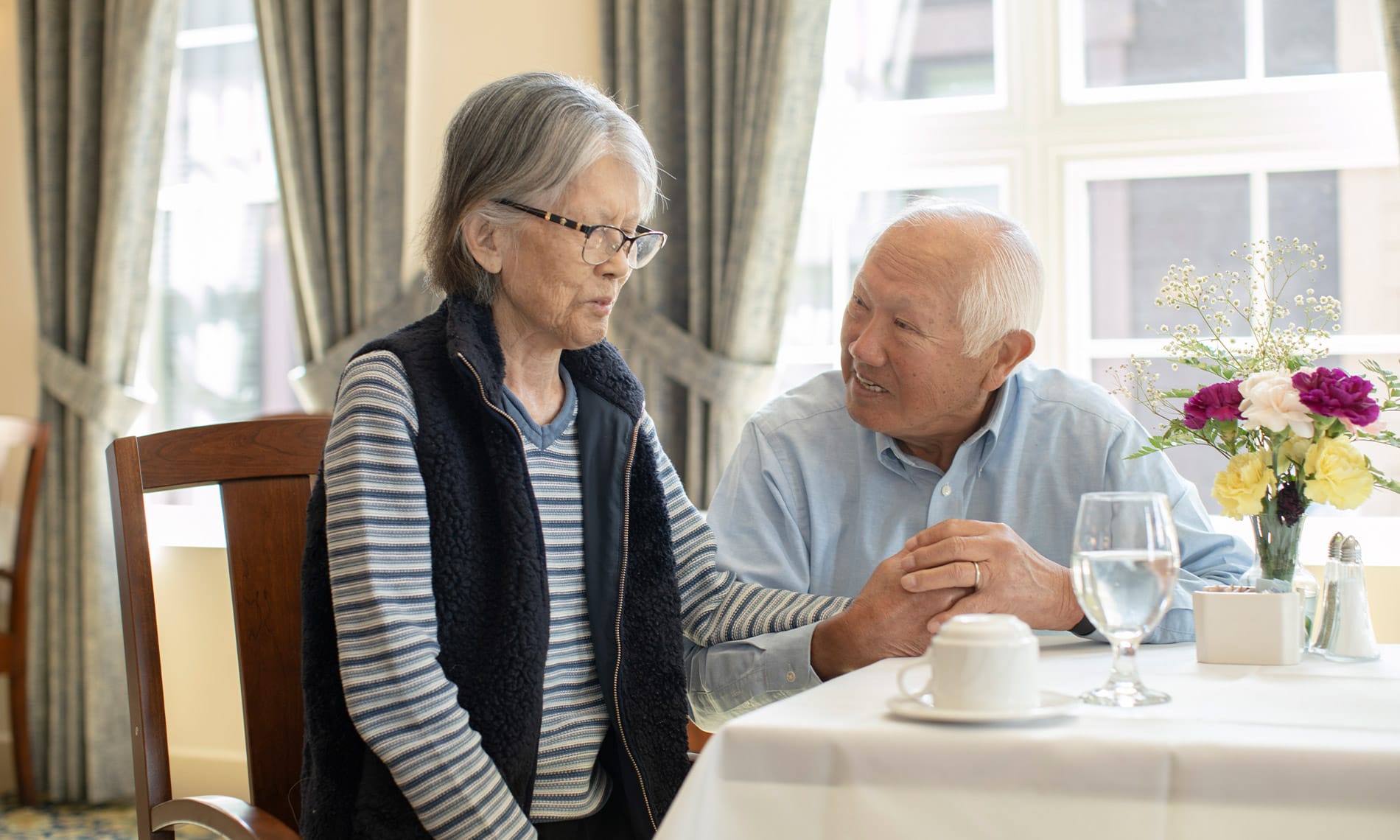 An elderly gentleman and his wife holding hands while enjoying a meal together as part of the assisted living program at the Fountains at the Albemarle