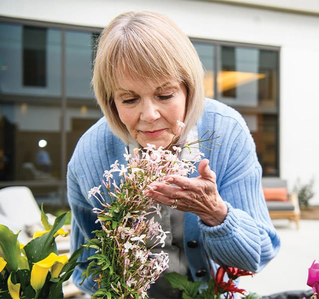 A woman in a blue sweater leaning over to smell light pink flowers in the garden