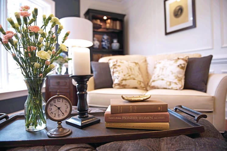 A coffee table set with a vintage clock, candle stick and several books, a couch in the midground with a grandfather clock in the background