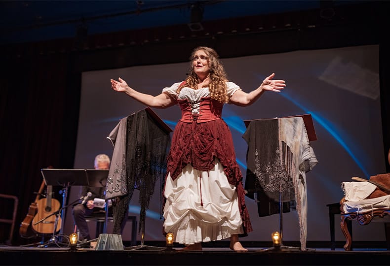 The Fountains at the Albemarle The Neighborhood - Tar River Players Female performer on the stage. She is dressed in period costume. Stage interior of Theatre during performance at night.