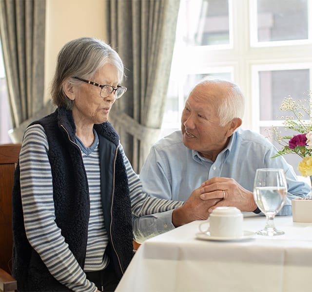 The Fountains at Albemarle Assisted Living An elderly gentleman and his wife holding hands while enjoying a meal together as part of the assisted living program at the Fountains at the Albemarle
