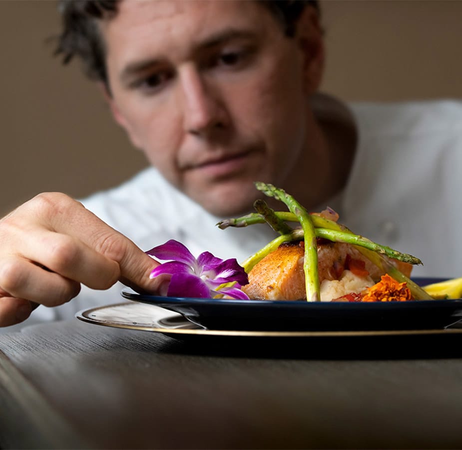 A chef placing a decorative flower on the edge of a meal to enhance the presentation A chef placing a decorative flower on the edge of a meal to enhance the presentation